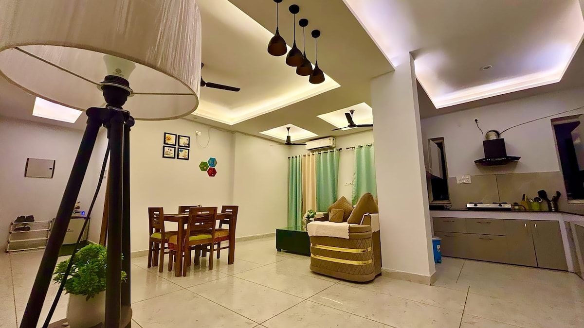 A spacious living area features a dining table with four chairs and a cozy seating arrangement including a sofa. Natural light enters through large windows, highlighting the calming color scheme. Modern kitchen amenities are visible in the background, complemented by ambient ceiling lighting.