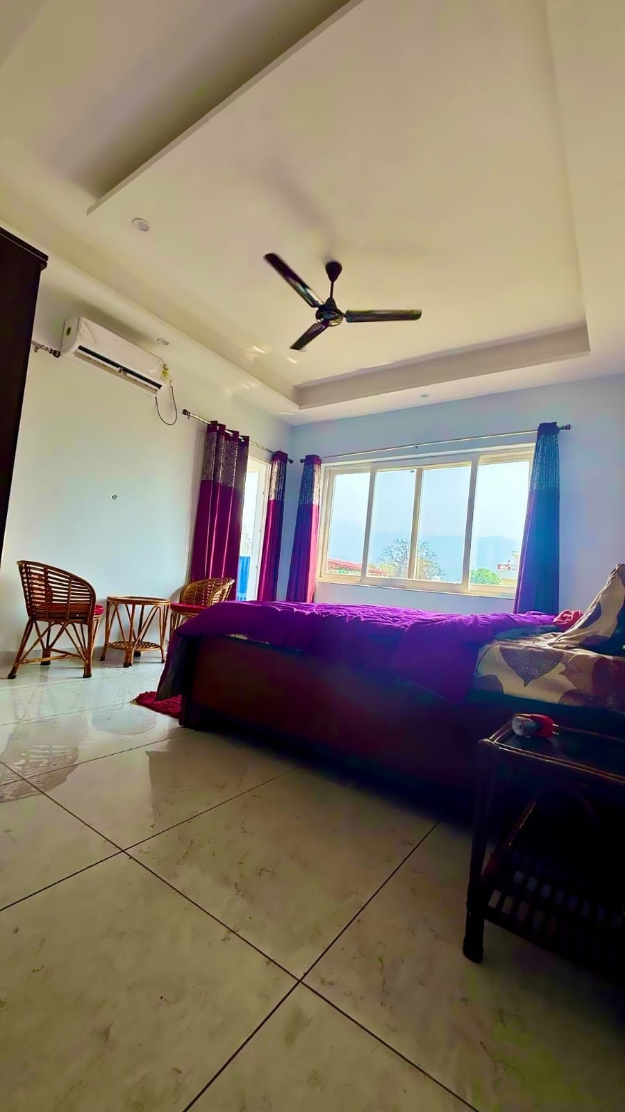 A bright bedroom is showcased with large glass windows offering mountain views. The space includes a spacious bed, two wicker chairs, and a dark wooden furniture set. Soft sunlight illuminates the room, while a ceiling fan adds comfort.
