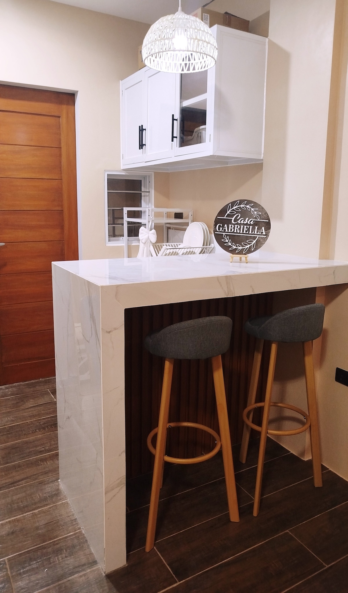 A modern bar area features a sleek white countertop with minimalist decor above. Two upholstered stool chairs are placed at the counter, and the wooden paneling adds warmth. A stylish pendant light hangs overhead, enhancing the contemporary feel of the space.