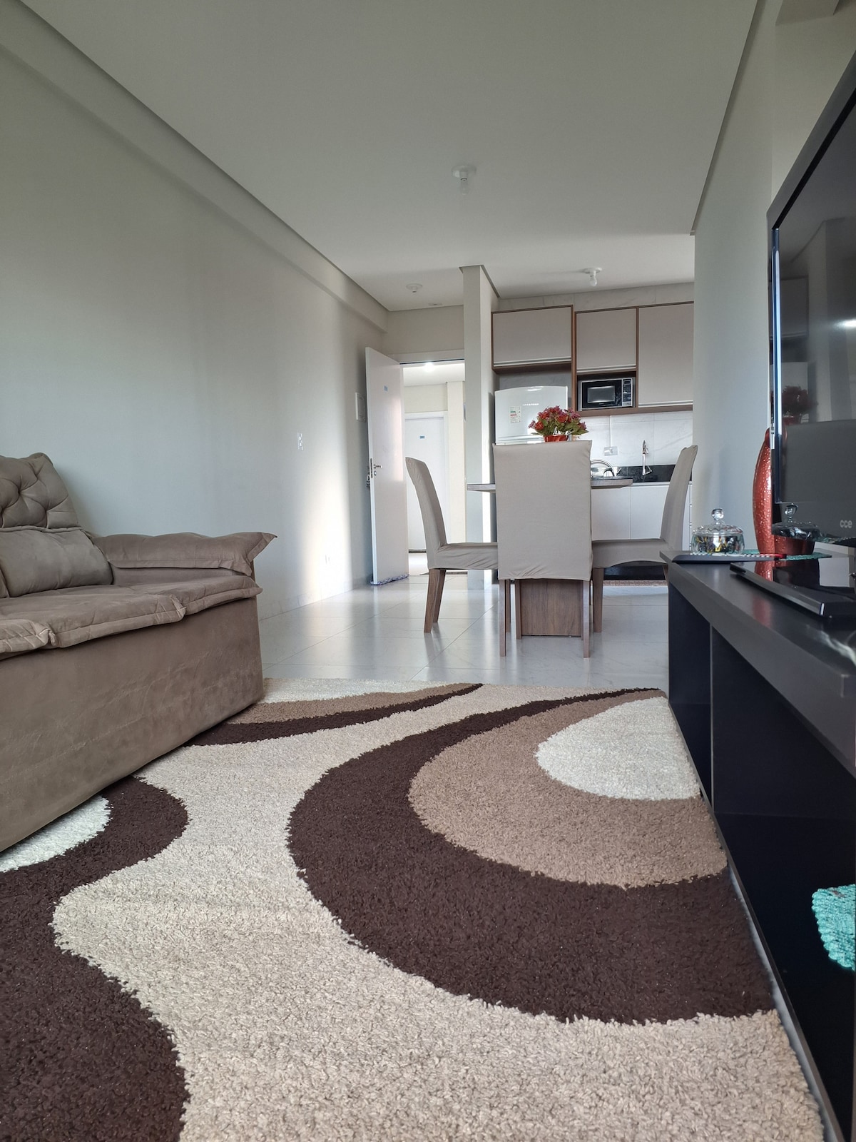 A well-lit living space is shown, featuring a comfortable brown sofa positioned near a large television. A dining table with chairs is visible in the background, while a soft, patterned rug adds texture to the floor. The kitchen area is partially seen through an open layout.