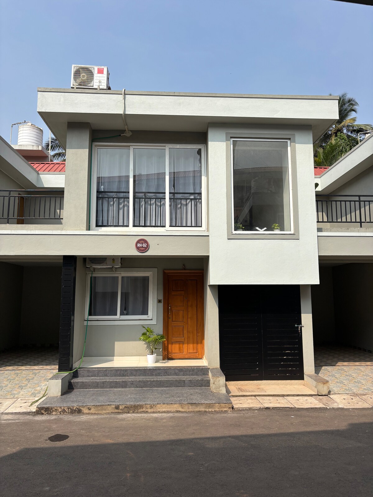 The villa's exterior features a modern design with clean lines. A wooden door is complemented by large windows that provide natural light. A small potted plant is placed at the entrance, and the facade includes a mix of neutral tones, adding to the contemporary appearance.