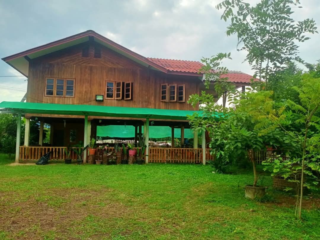 A wooden house with a red-tiled roof is set in a spacious green yard. The structure features multiple windows and a covered porch, surrounded by lush trees and plants. The area is tranquil, offering a serene retreat in nature.
