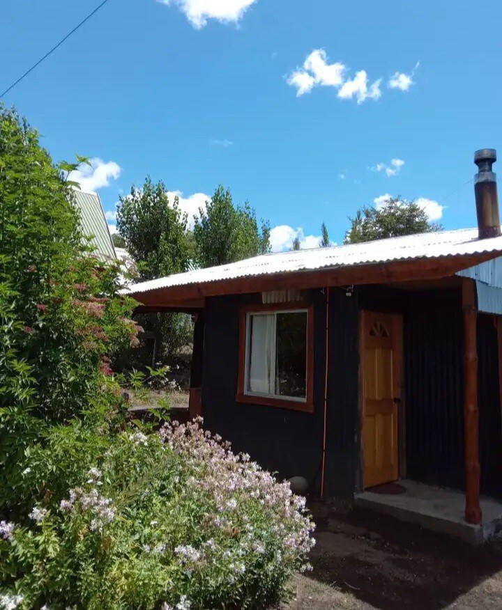 The exterior of the cabin features a charming wooden door framed by greenery, with colorful flowers in the foreground. The structure is topped with a sloped roof and includes a window that allows natural light to enter the interior. A clear blue sky is visible above.