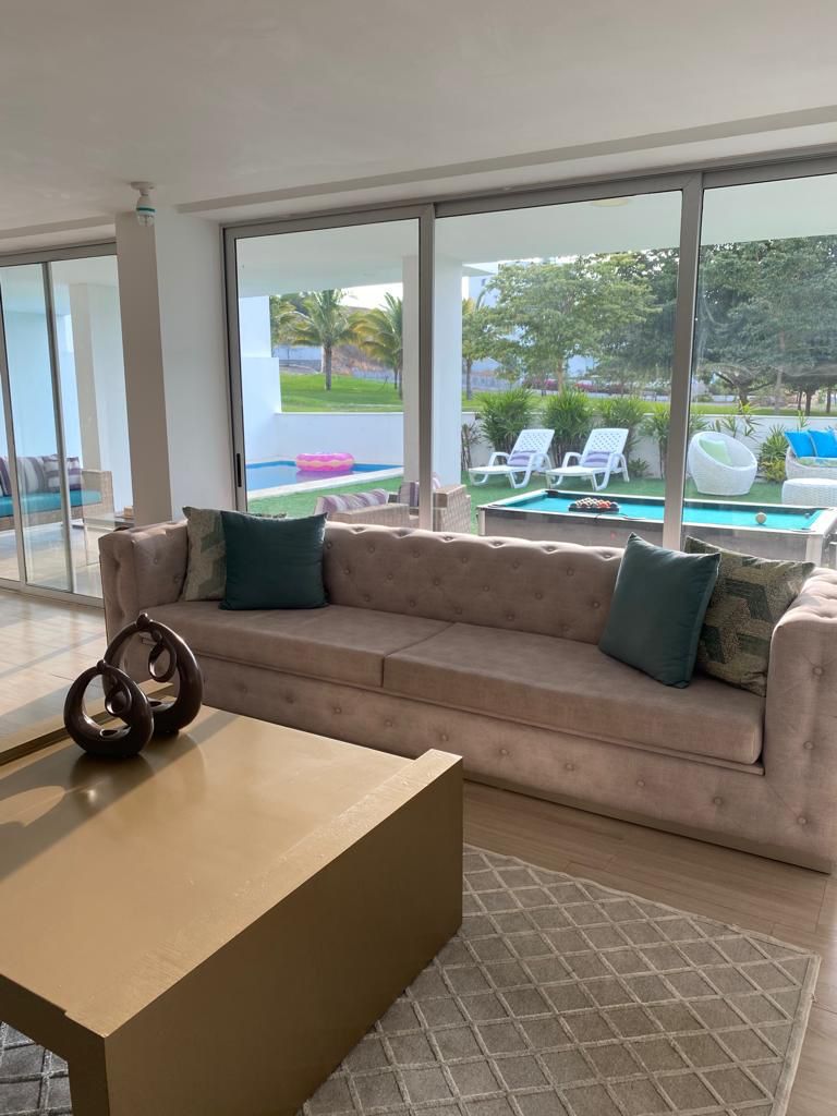 A spacious living area features a plush, tufted sofa adorned with decorative pillows. Large glass doors offer a view of the outdoor space, which includes a pool surrounded by tropical greenery. A coffee table sits on a textured rug, adding warmth to the space.