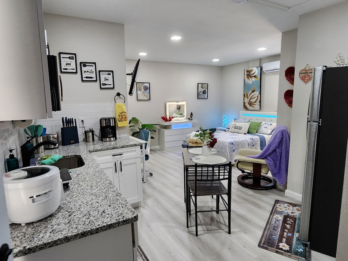 A modern studio apartment is shown, featuring a well-equipped kitchen with white cabinetry and granite countertops. An inviting seating area includes a small table and chairs. The sleeping area features a cozy bed, with colorful bedding and decorative elements enhancing the style.