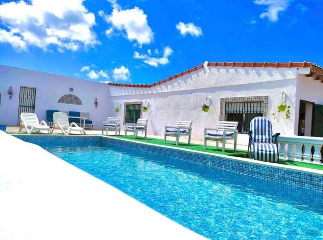 A sparkling private pool is framed by a spacious terrace, featuring multiple sun loungers for relaxation. The villa's white exterior contrasts against the vibrant blue sky, with green accents from hanging plants enhancing the setting. Large windows allow natural light to brighten the area.