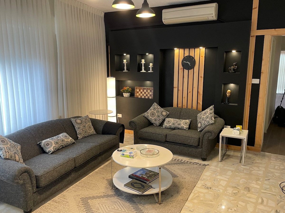 A modern living area is showcased, featuring two sofas adorned with patterned cushions. A round coffee table sits in the center, accompanied by a side table. Wall shelves display decorative items, while soft lighting enhances the space's comfort.