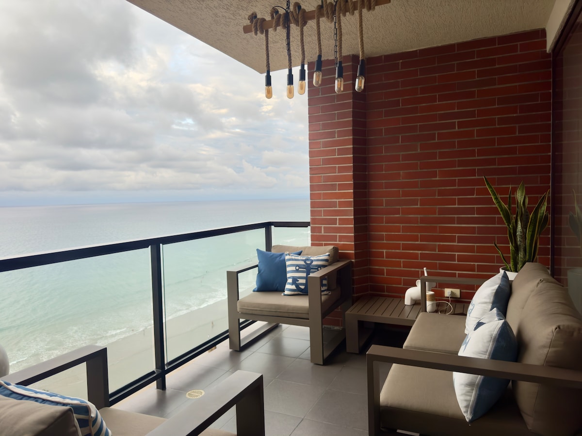A balcony offers a serene view of the ocean, featuring two comfortable lounge chairs with striped pillows. Soft lighting fixtures hang above, and a potted plant adds a touch of greenery. The glass railing provides an unobstructed view of the shoreline.