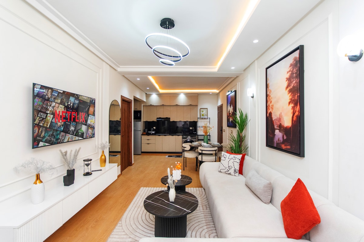 A modern living area is equipped with a 55-inch television displaying Netflix. A light-colored sofa is adorned with red and patterned cushions. Subtle lighting fixtures and wall art enhance the ambiance. A dining area is visible in the background, alongside a well-equipped kitchenette.