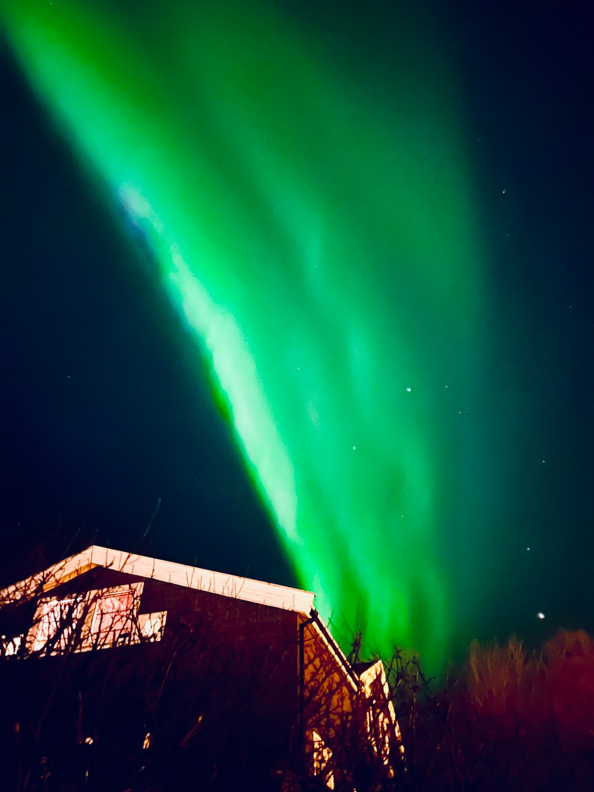 Northern lights illuminate the night sky with vibrant green hues, cascading above the roof of the nearby house. A backdrop of stars adds depth to the scene, creating a serene and captivating atmosphere in the remote location.