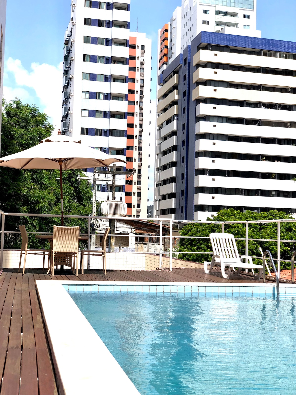 A rooftop pool is framed by a wooden deck, featuring a sun umbrella and lounge chairs. Surrounding high-rise buildings provide a contrasting backdrop against the clear blue sky, contributing to a relaxing outdoor atmosphere.