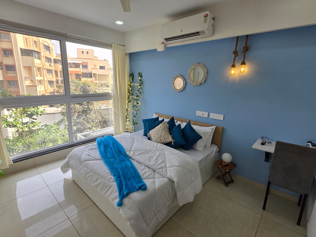 A modern studio bedroom features a spacious bed dressed in white linens and accentuated with blue and turquoise cushions. A small work desk is positioned beside the bed, and natural light floods the room through large windows adorned with light curtains.