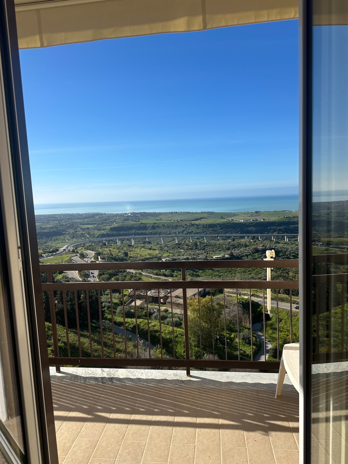 A spacious balcony is visible, offering a sweeping view of the coastline and lush landscape. A clear blue sky complements the scene, while a comfortable chair provides a spot for relaxation. The balcony is partially shaded by a wide awning.