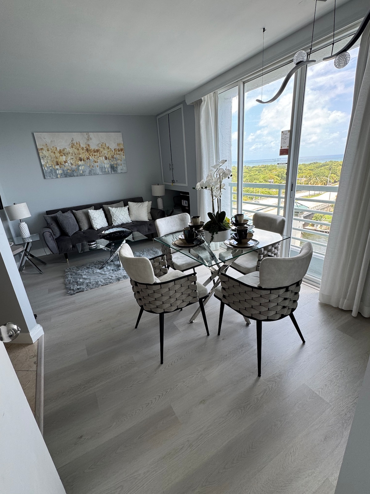 A modern living area features a sectional sofa adorned with decorative pillows, complemented by a glass dining table surrounded by stylish chairs. Large windows allow natural light to brighten the space, highlighting the neutral color palette and a piece of artwork on the wall.