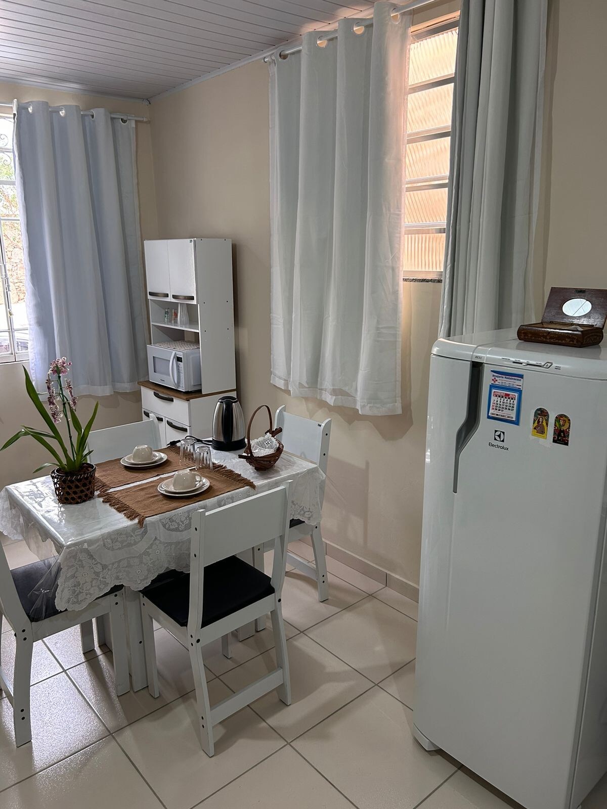 A dining area features a small table set for two, complemented by white chairs and a placemat. A kettle and decorative items are arranged on the table. Nearby, a white refrigerator stands adjacent to a small shelf with kitchen appliances and a potted orchid adds a touch of greenery.
