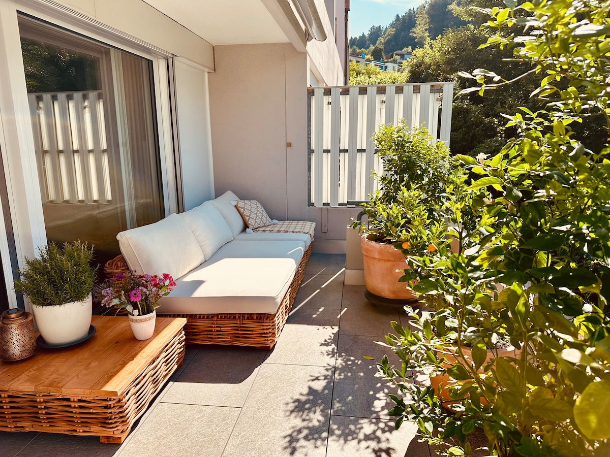 A comfortable terrace area is shown, featuring a spacious outdoor seating arrangement with light-colored cushions. Potted plants and flowers add greenery, while a surrounding barrier offers privacy. Sunlight illuminates the space, enhancing the tranquil atmosphere of this outdoor setting.