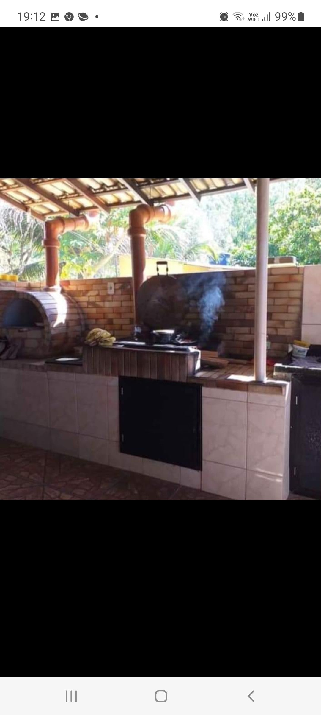 An outdoor kitchen area is presented, featuring two traditional clay ovens. A stone countertop is visible, with cooking utensils and pots placed neatly. Sunlight filters through the open structure, highlighting the textured walls and surrounding greenery.