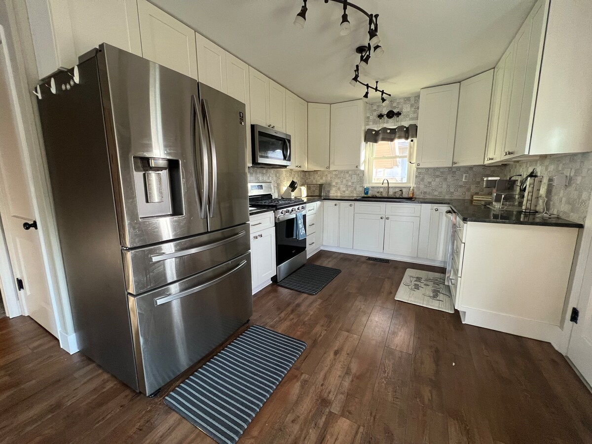 A modern kitchen features stainless steel appliances, including a large refrigerator and a gas stove. White cabinetry surrounds the space, complemented by a neutral tile backsplash. The flooring is wood, and two textured mats are placed near the stainless steel sink and stove.
