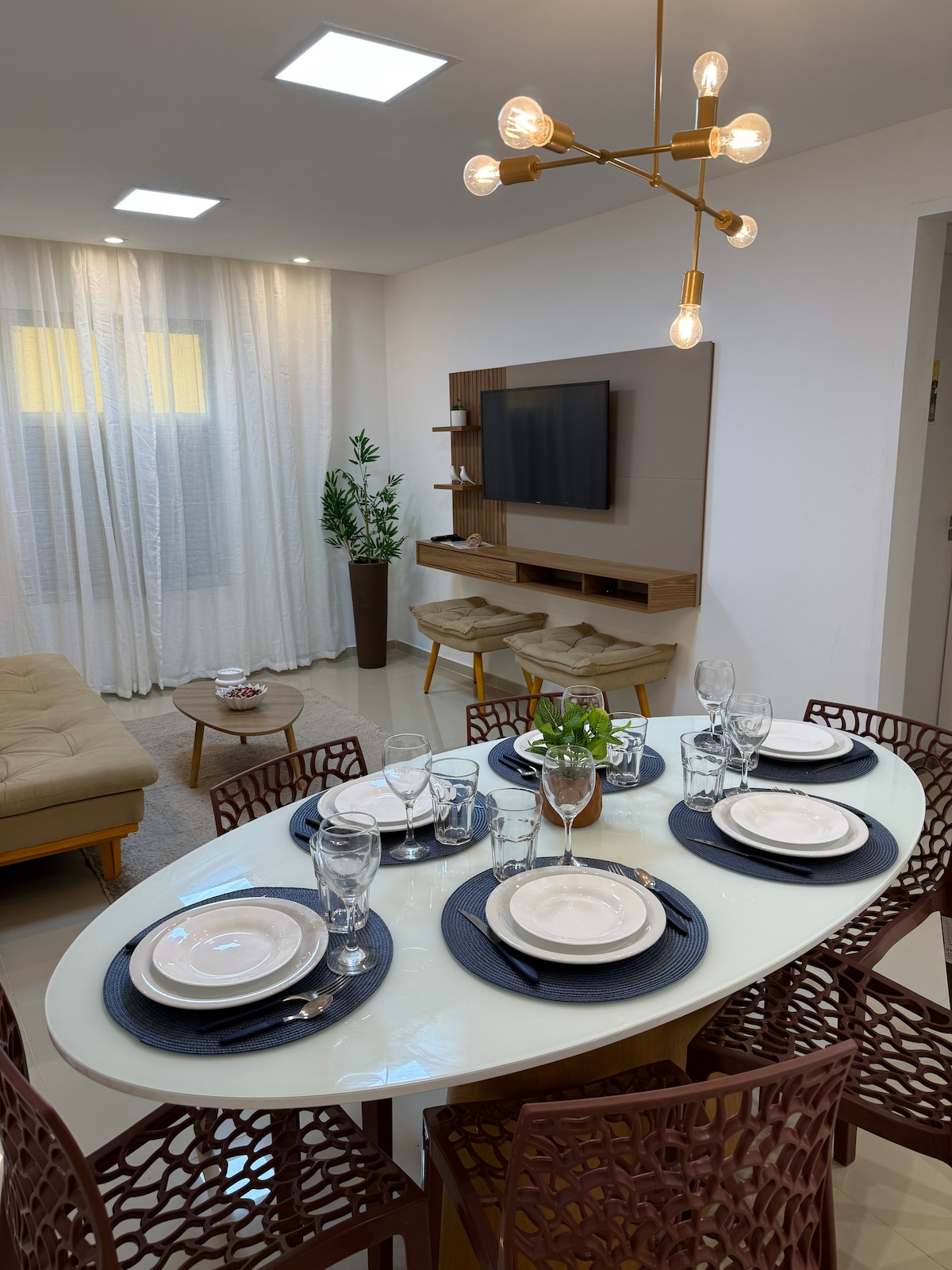 A modern dining area features an oval table set with plates, glasses, and utensils. Elegant lighting fixtures hang overhead, illuminating the space. A cozy seating area with a light-colored couch and a wall-mounted TV is visible in the background, complemented by decorative plants.
