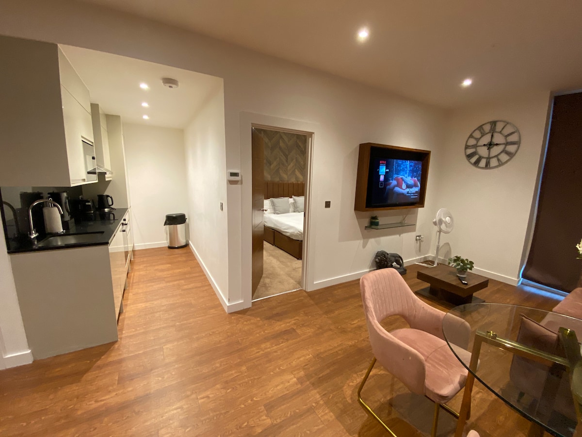 An open-plan space is visible, featuring a modern kitchen with sleek cabinetry and appliances. A dining area with a glass table and pink chairs is present, alongside a living area equipped with a wall-mounted television. A bedroom with a spacious bed can be glimpsed through an open doorway.
