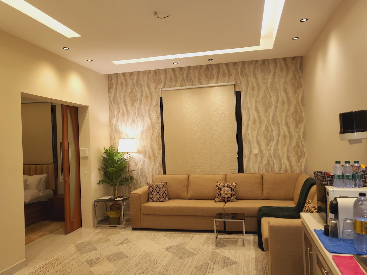 A spacious living area features a long, comfortable sofa with decorative cushions, accompanied by a sleek coffee table. A subtle plant adds a touch of greenery. The area is illuminated by soft ceiling lights, and a large window is covered by a textured shade.