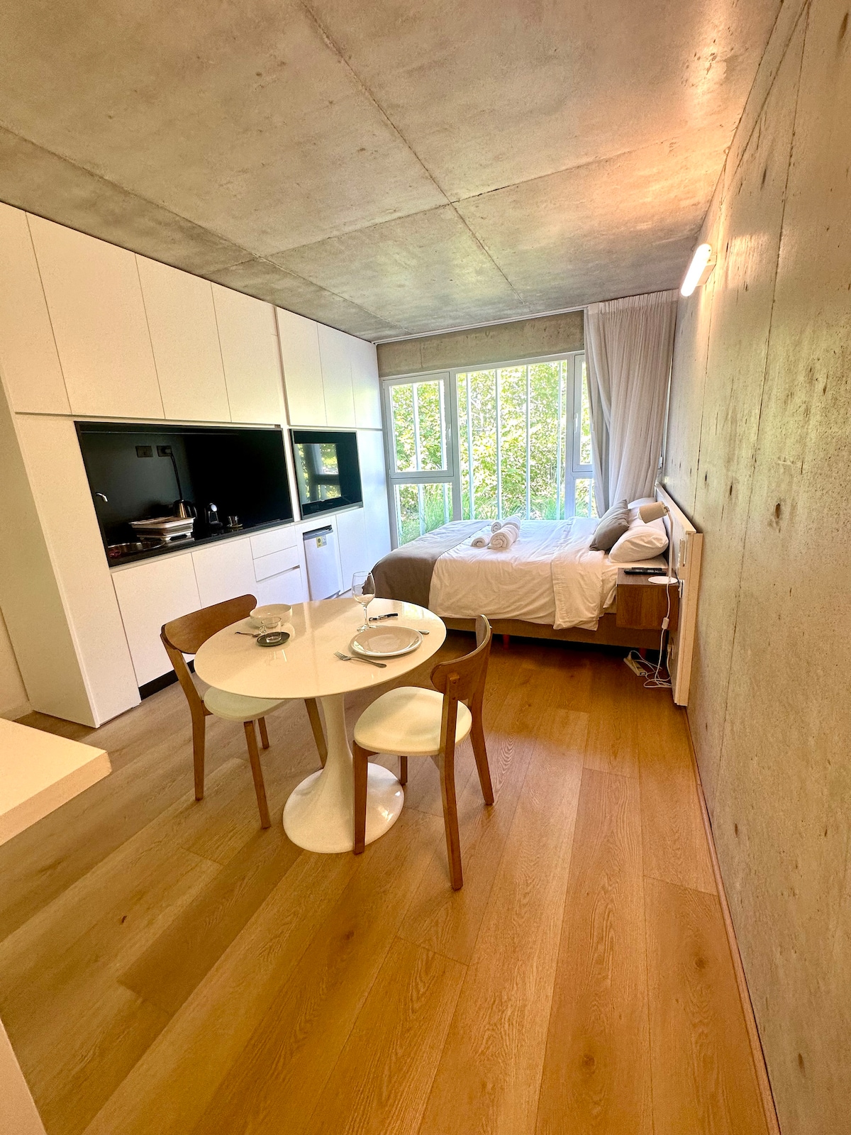 A modern studio space is shown, featuring a queen bed against a light-colored wall and large windows providing ample natural light. A compact dining table with two chairs is positioned near the kitchen area, enhancing functionality within the open layout.