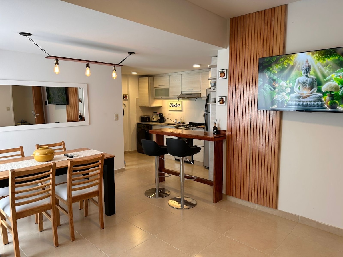 An open-plan living space displays a dining area with a wooden table surrounded by six chairs. A modern kitchen with light cabinetry and stainless-steel appliances is visible. A wall-mounted television features a serene image of a Buddha. Natural light enhances the neutral color palette.
