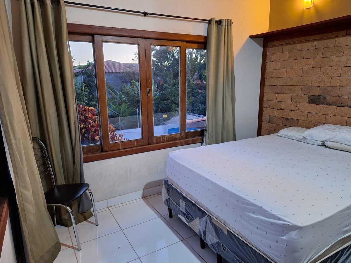 A well-lit bedroom is displayed featuring a spacious bed with a white mattress and bedding. A wooden accent wall complements a large window with views of the outdoors. A simple chair is positioned nearby, and light-toned curtains frame the window.