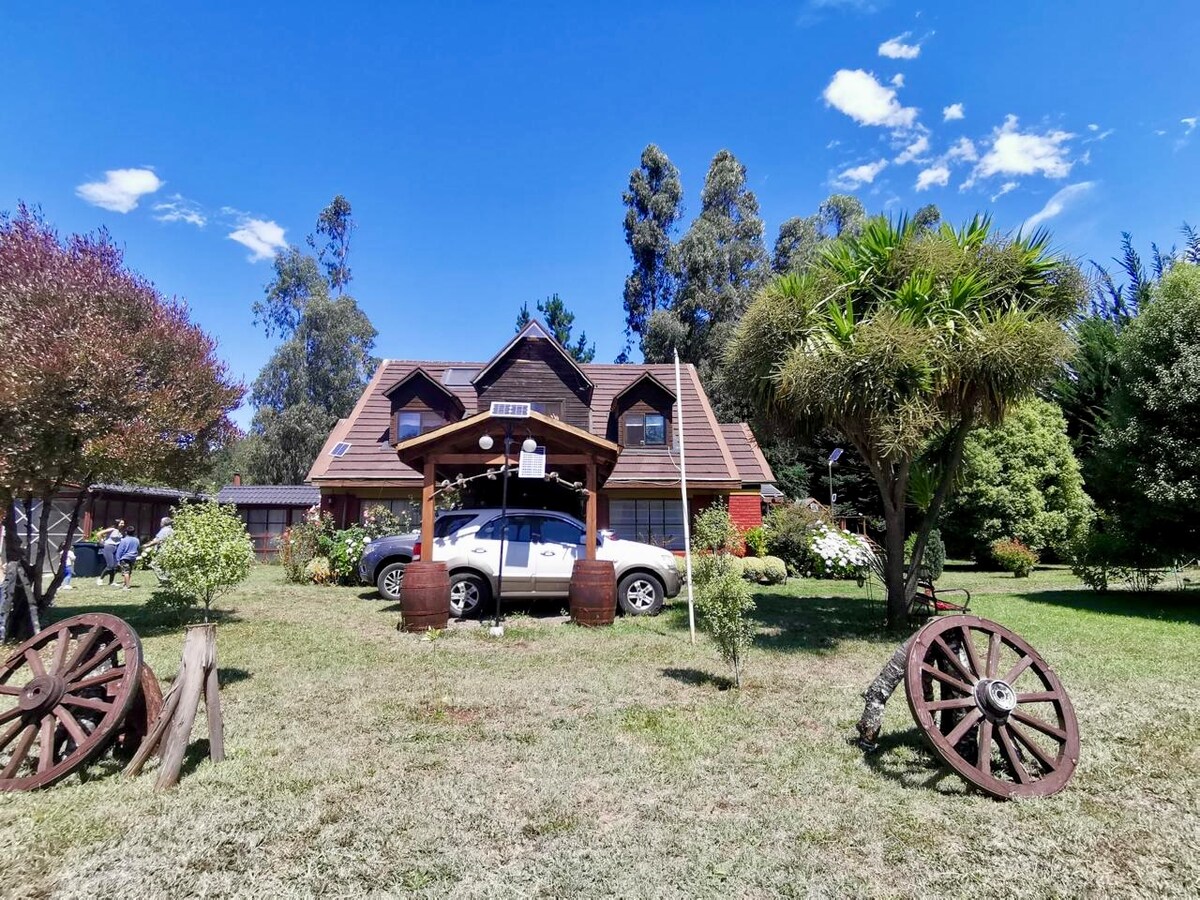A charming country house emerges amidst lush greenery, featuring a sloped roof and spacious wooden porch. Two vehicles are parked beneath a shaded area, surrounded by vibrant garden beds and tall trees. Decorative wagon wheels enhance the inviting exterior.