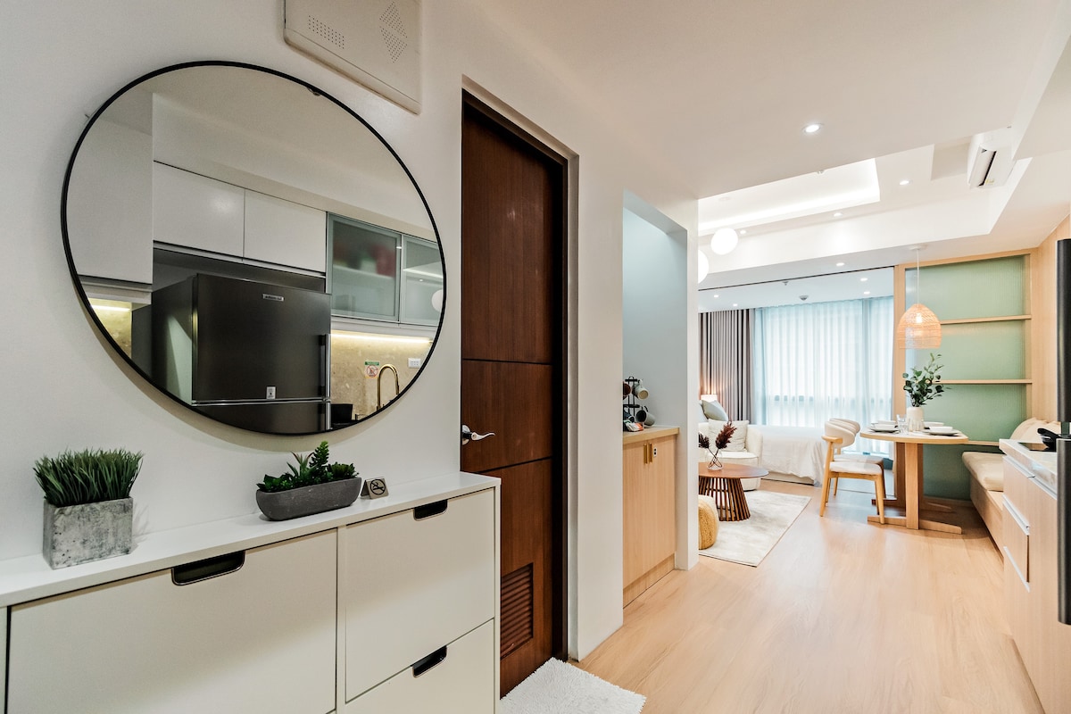 A circular mirror reflects a modern entryway featuring sleek cabinetry and a small potted plant. The open layout reveals a living area in soft natural light, with a dining table and comfortable seating visible in the background.