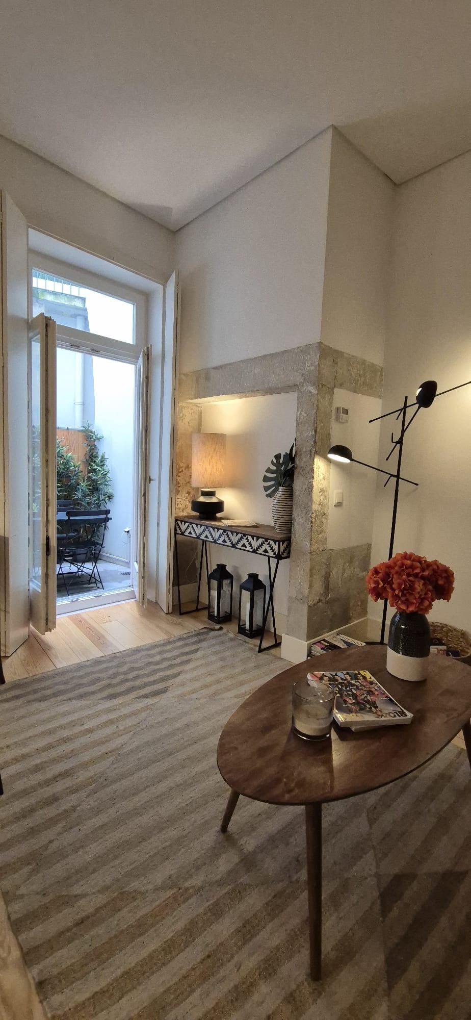 A bright corner of the loft features large glass doors opening to a small outdoor terrace. A round wooden table is set with a decorative vase of flowers, and modern lighting fixtures enhance the space. A patterned rug covers the wooden floor, providing a warm touch.
