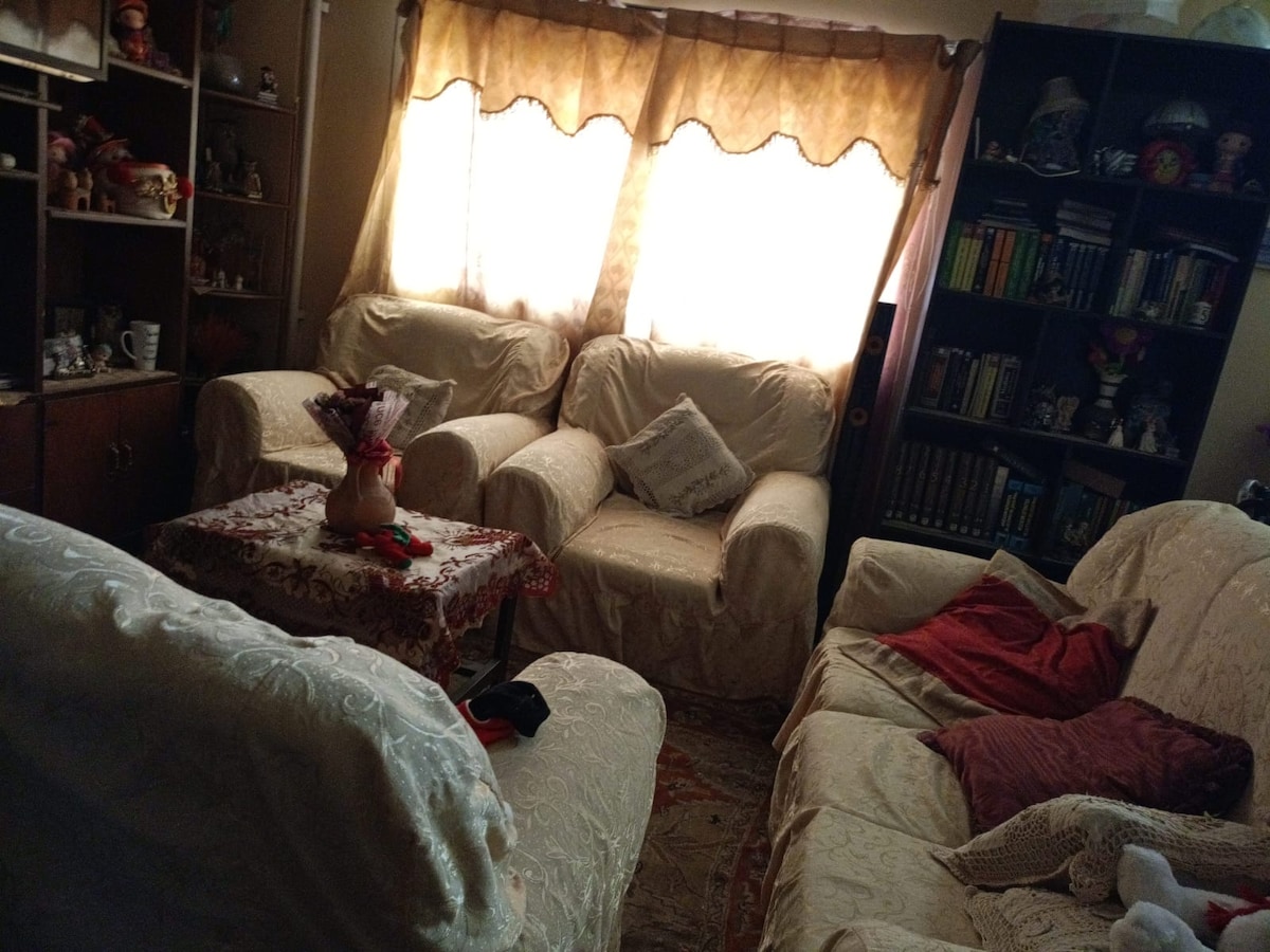 A cozy living area is furnished with a set of light-colored sofas and a small coffee table. Natural light enters through two large windows, partially covered by sheer curtains. Bookshelves and decorative items add a personal touch to the space.