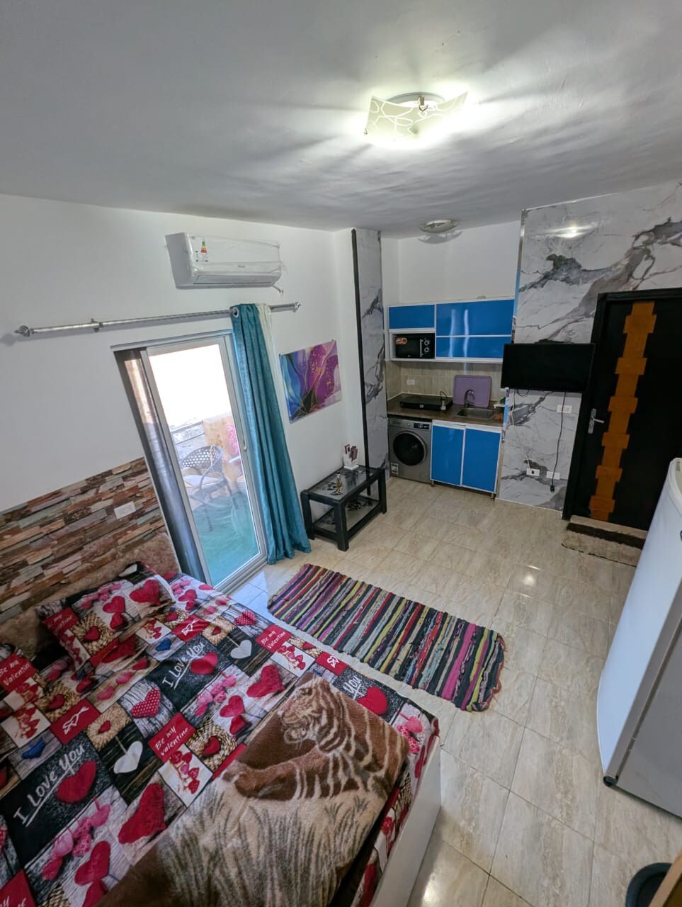 A compact living space features a spacious bed dressed with a colorful patterned comforter. A well-equipped kitchenette is visible, set against a marbled backdrop. Natural light enters through a window adorned with light blue curtains, illuminating the space designed for comfort and functionality.