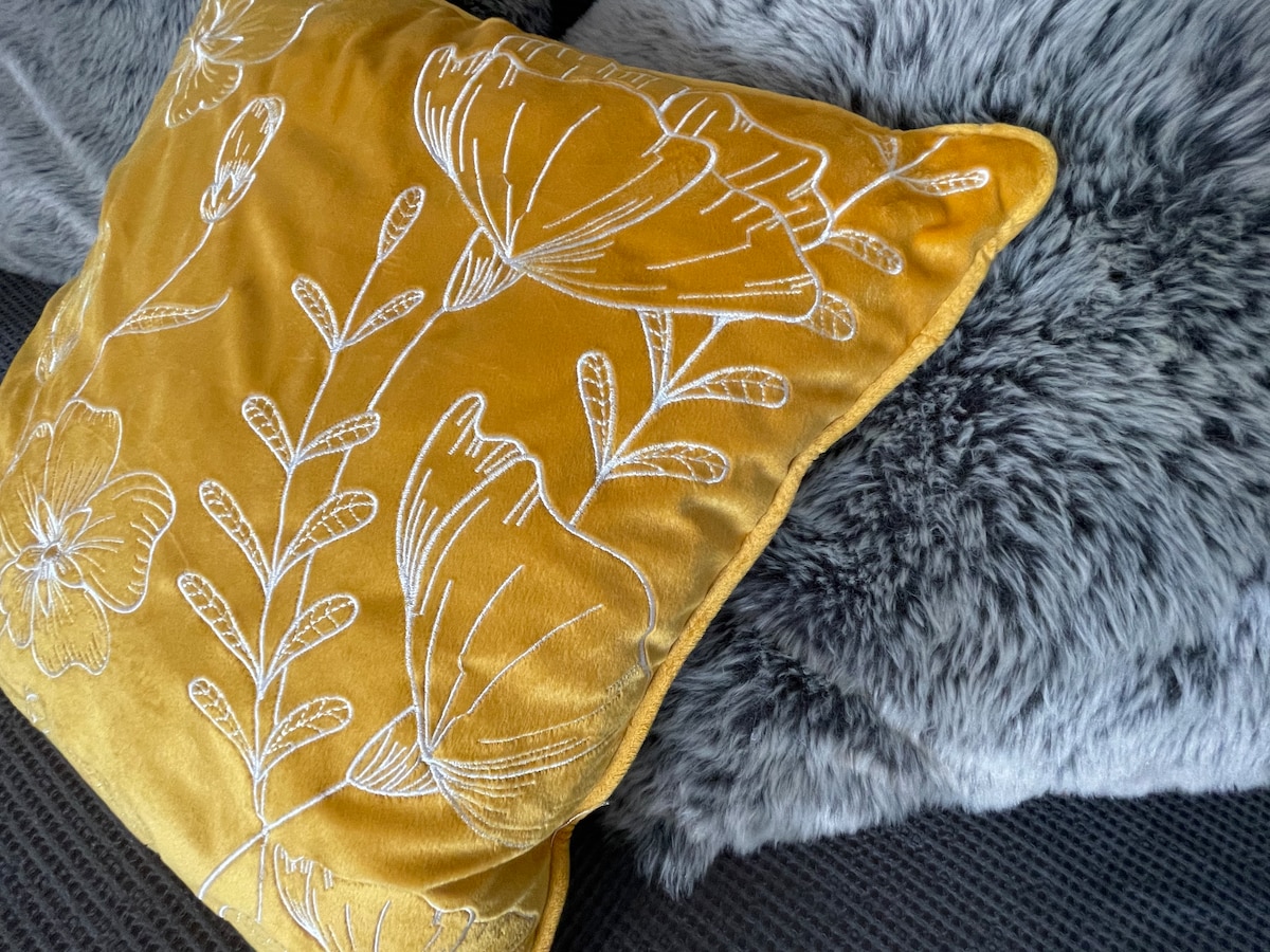 A textured yellow pillow with floral embroidery is positioned alongside a soft, grey faux-fur pillow. The arrangement enhances the cozy appearance of the seating area, inviting comfort and relaxation.