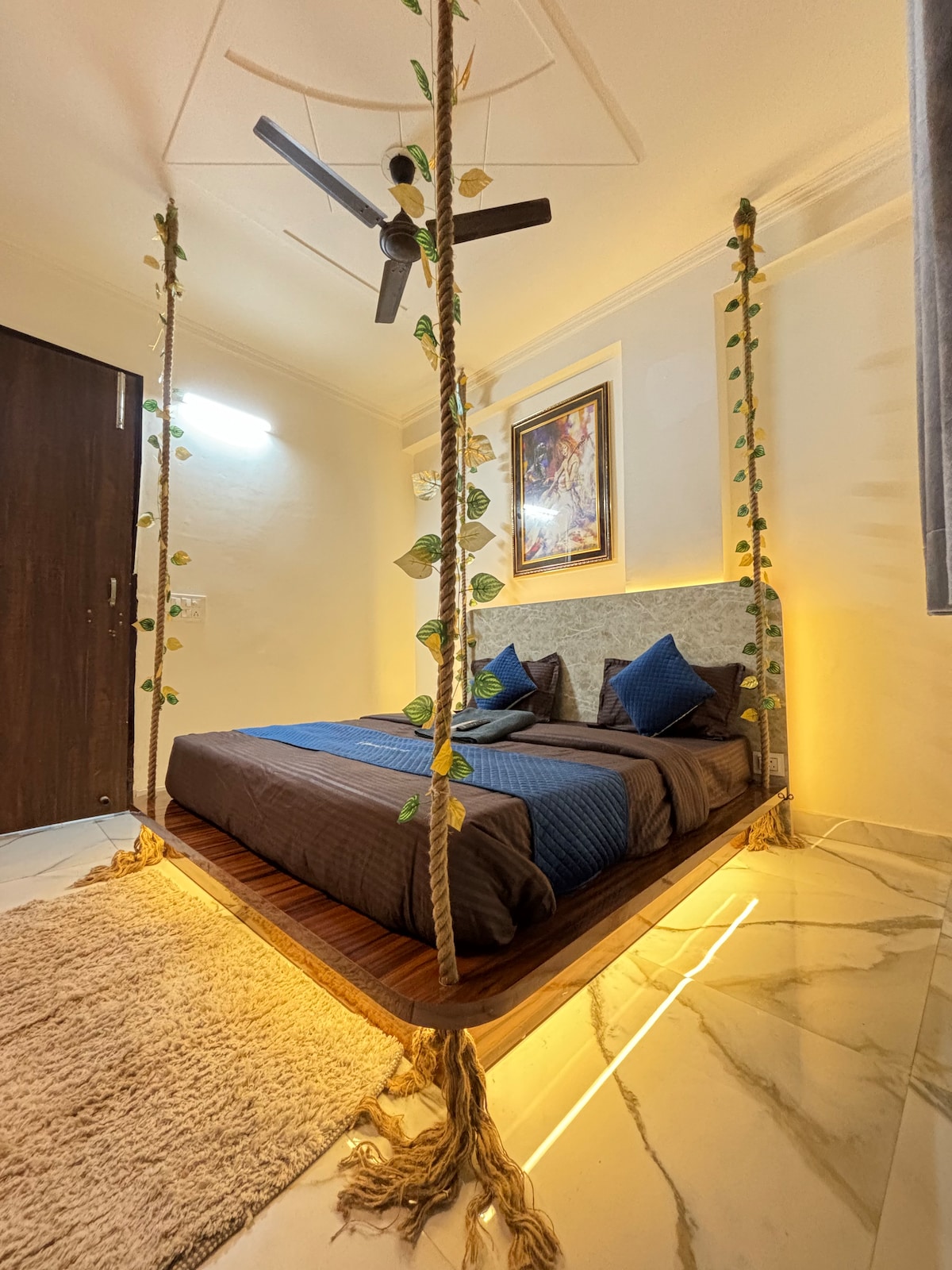 A unique sleeping area features a suspended bed, framed with decorative ropes and greenery. Soft lighting casts a warm glow on the space, which is complemented by a large piece of artwork above the bed and plush carpeting underfoot.