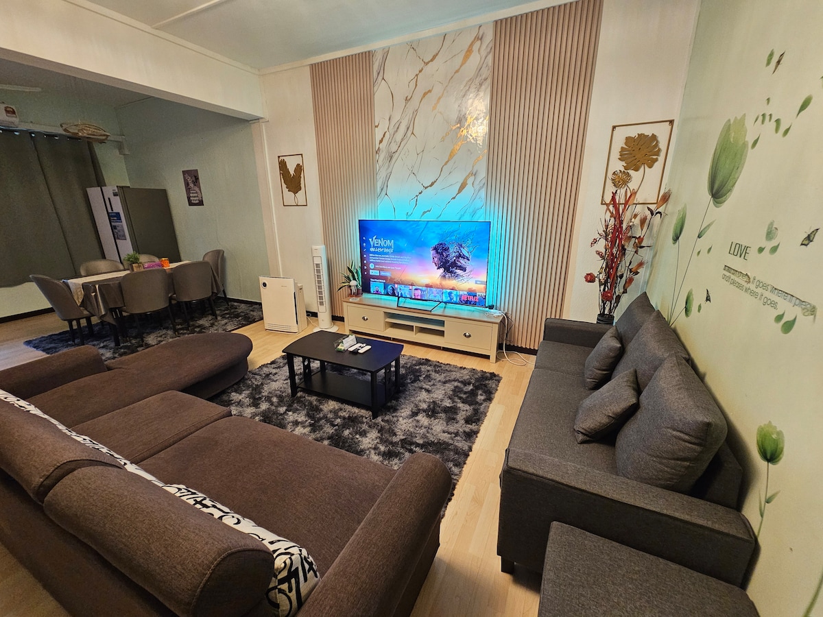 A living area is presented with two sectional sofas facing a coffee table. A large flat-screen television is mounted in front, with a decorative wall featuring floral designs. The space is illuminated by natural light from the windows, creating a welcoming environment.