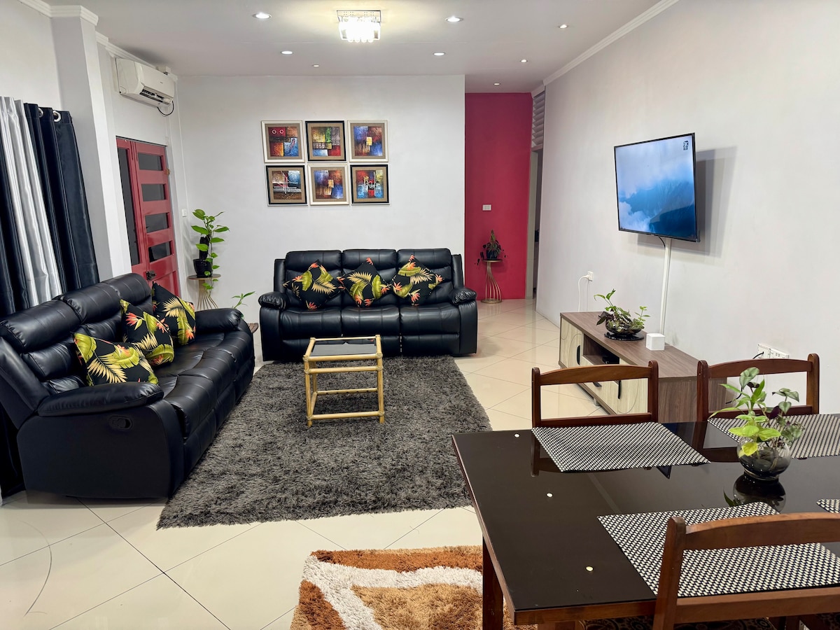 A spacious living area is highlighted by two black leather sofas adorned with decorative cushions. A dark rug sits beneath a glass coffee table, complemented by an indoor plant. A wooden dining table with light-colored chairs is positioned nearby, along with a wall-mounted television and vibrant wall art.