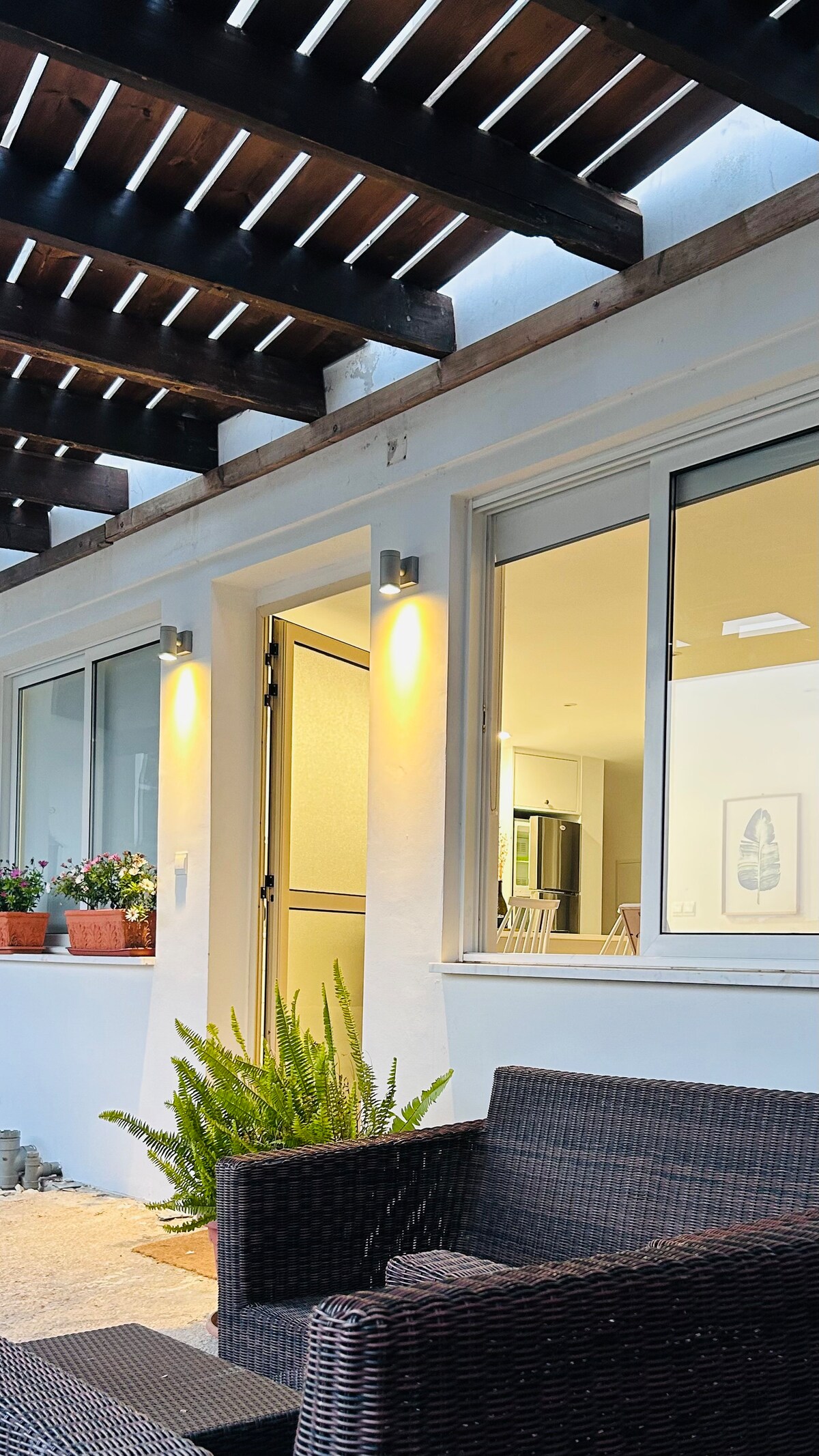 A contemporary outdoor seating area is visible, featuring a dark wicker chair and small table. Potted plants add greenery, and soft lighting enhances the adjacent entrance. Through the glass door, a glimpse of the modern kitchen and dining area can be seen, creating an inviting atmosphere.