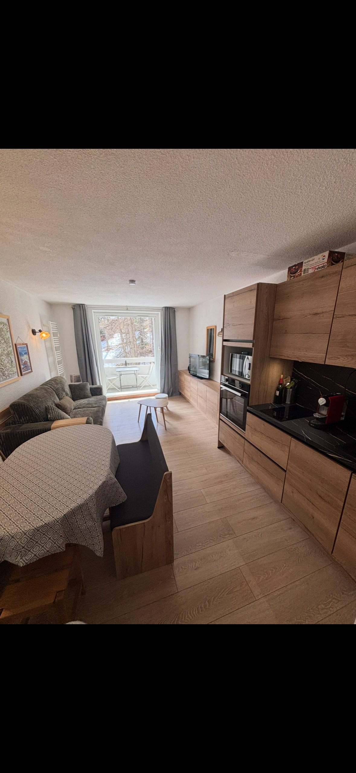 A light-filled living space features a cozy seating area with a couch beside a flat-screen television. A round dining table is positioned near a fully equipped kitchen, which showcases modern appliances and ample cabinetry. Large glass doors lead to a balcony with views of the outdoors.