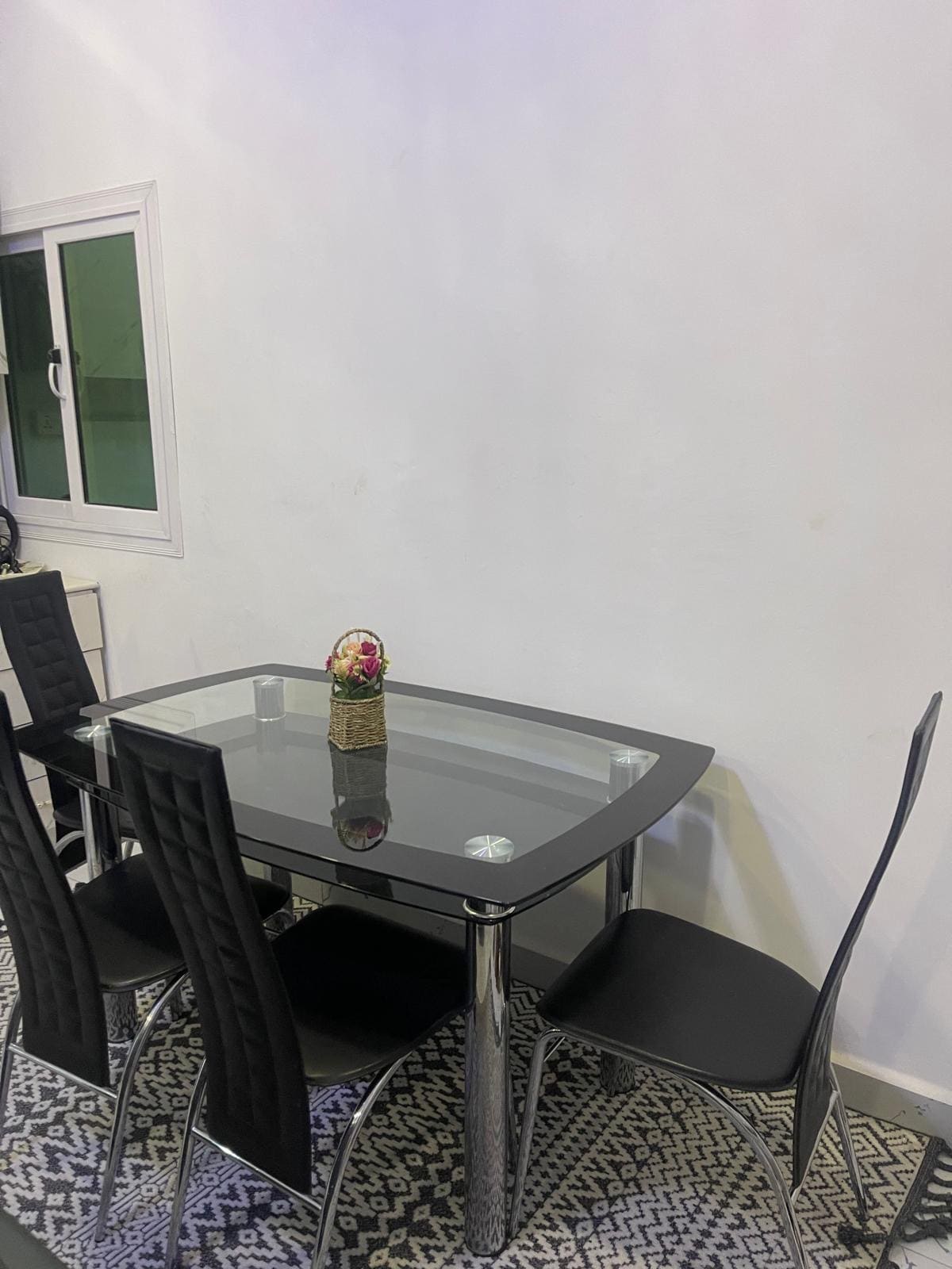 A dining area is presented with a glass-topped table surrounded by four black chairs. A small floral arrangement is placed at the center of the table. A patterned rug is visible beneath the furniture, and a window allows natural light into the space.
