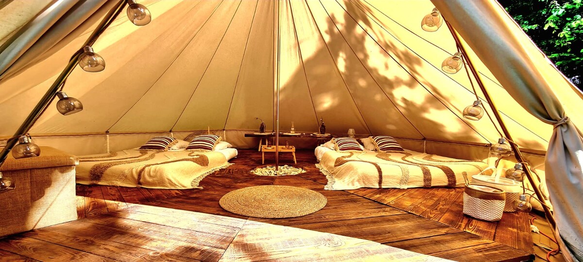 Inside the spacious tent, soft flooring is complemented by wooden surfaces. Two beds arranged with striped bedding and light throws are visible, alongside a central table adorned with decorative items. Subtle lighting is provided by hanging lamps, creating a comfortable and functional environment.