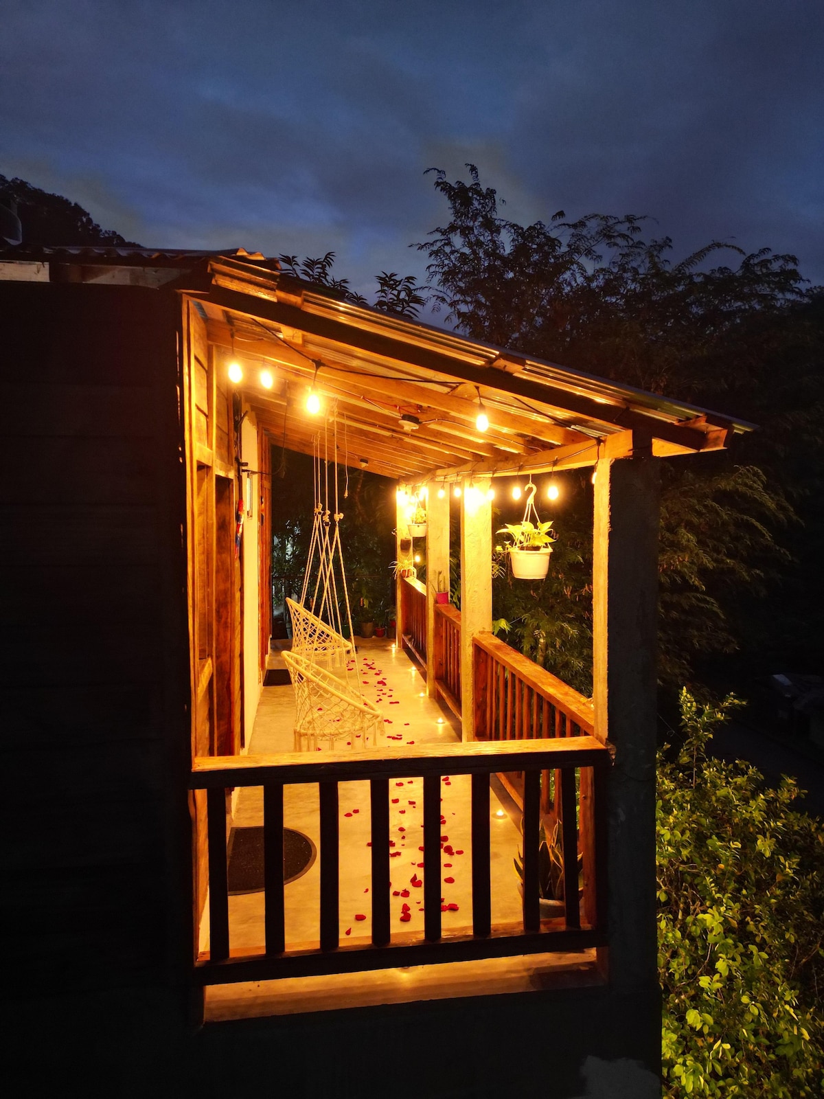 A warmly lit porch is framed by wooden railings and features a hanging swing. Soft lighting creates a cozy atmosphere, while scattered rose petals along the floor add a touch of charm. Lush greenery is visible in the background, enhancing the natural setting.