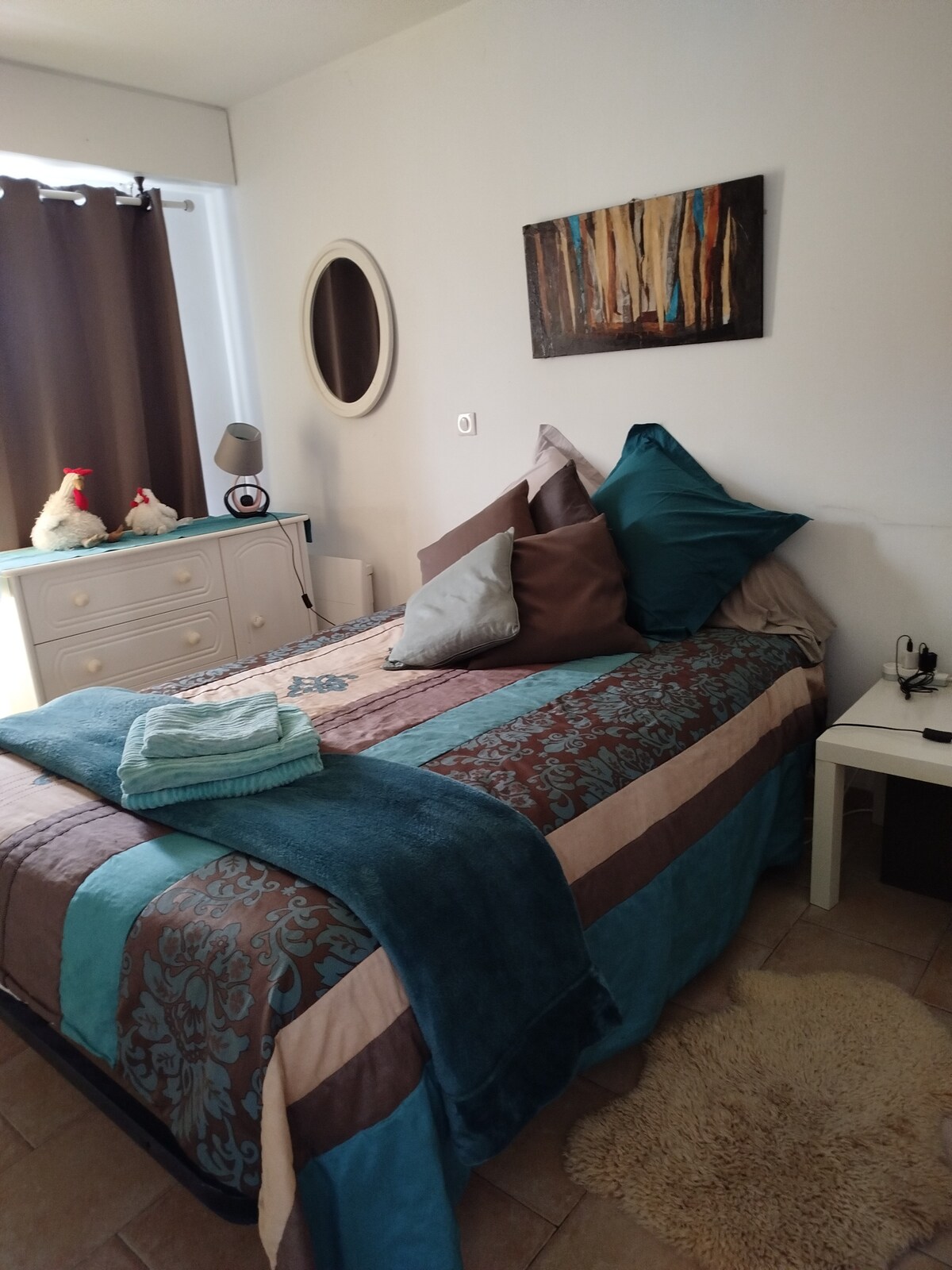 A comfortable bedroom features a spacious bed adorned with decorative pillows and a soft throw blanket. A small bedside table holds a lamp, and natural light enters through a window covered with dark curtains. A round mirror and artwork add character to the space.