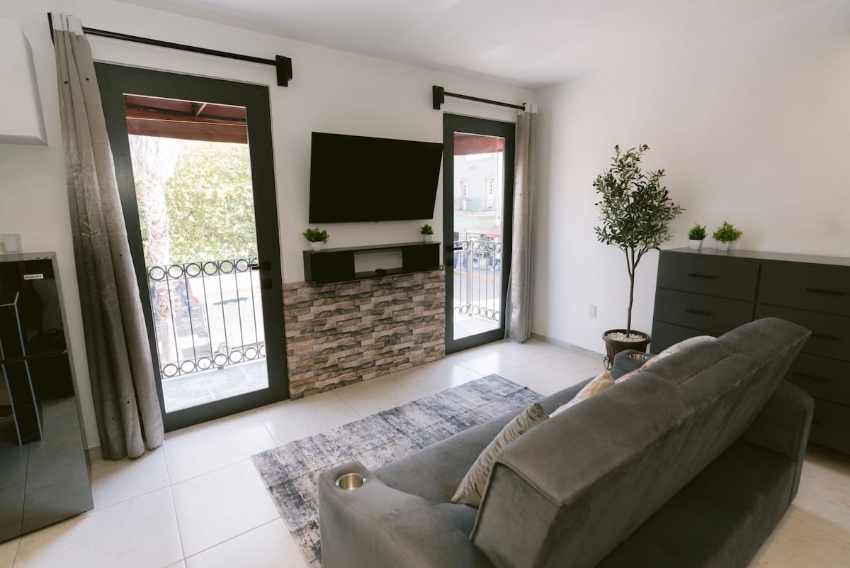 A light-filled living area features a comfortable gray sofa adjacent to a flat-screen TV mounted on a rustic stone panel. Two large windows allow natural light, framed by neutral curtains. A small potted plant decorates the space, adding a touch of greenery.