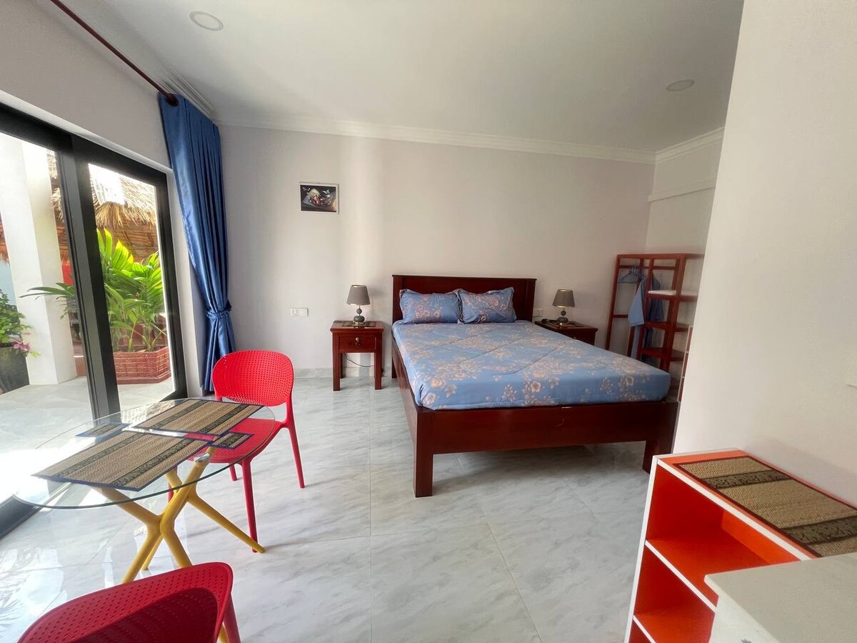 A bright and airy room is showcased, featuring a spacious bed with a floral blue bedspread. A small dining table with red chairs occupies one corner, while a wooden shelf and bedside tables provide additional functionality. Large glass doors open to a garden area, enhancing the natural light.