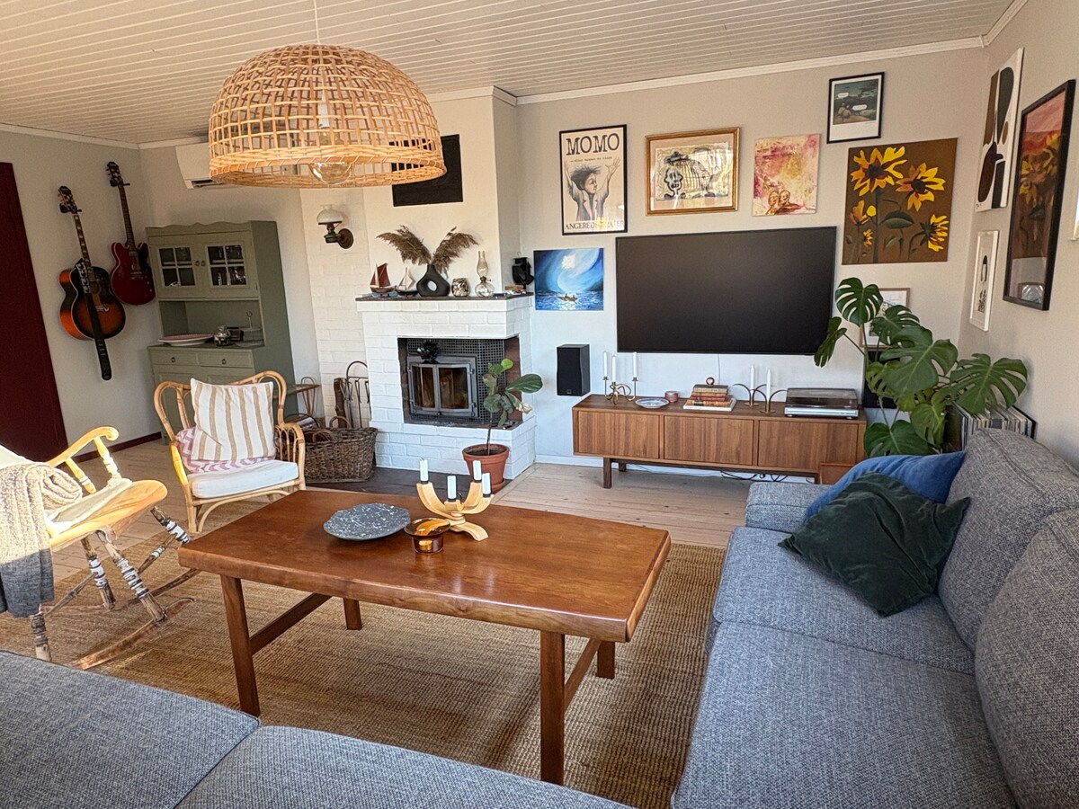 A comfortable living area features a mix of seating options, including a gray sectional sofa and stylish woven chairs. A coffee table sits at the center, complemented by decorative items. Wall art adds character, while a television is mounted above a low wooden cabinet.