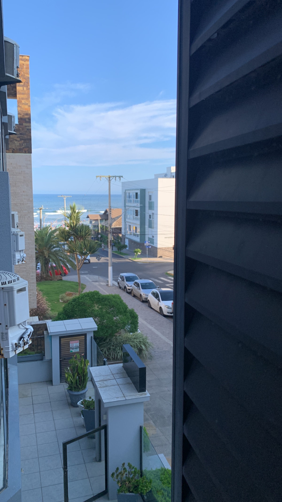 A view is captured from a balcony, showcasing the ocean visible in the distance. Lush greenery and a pathway lead towards the street, where parked cars are seen. Modern buildings line the road, under a clear blue sky with scattered clouds.