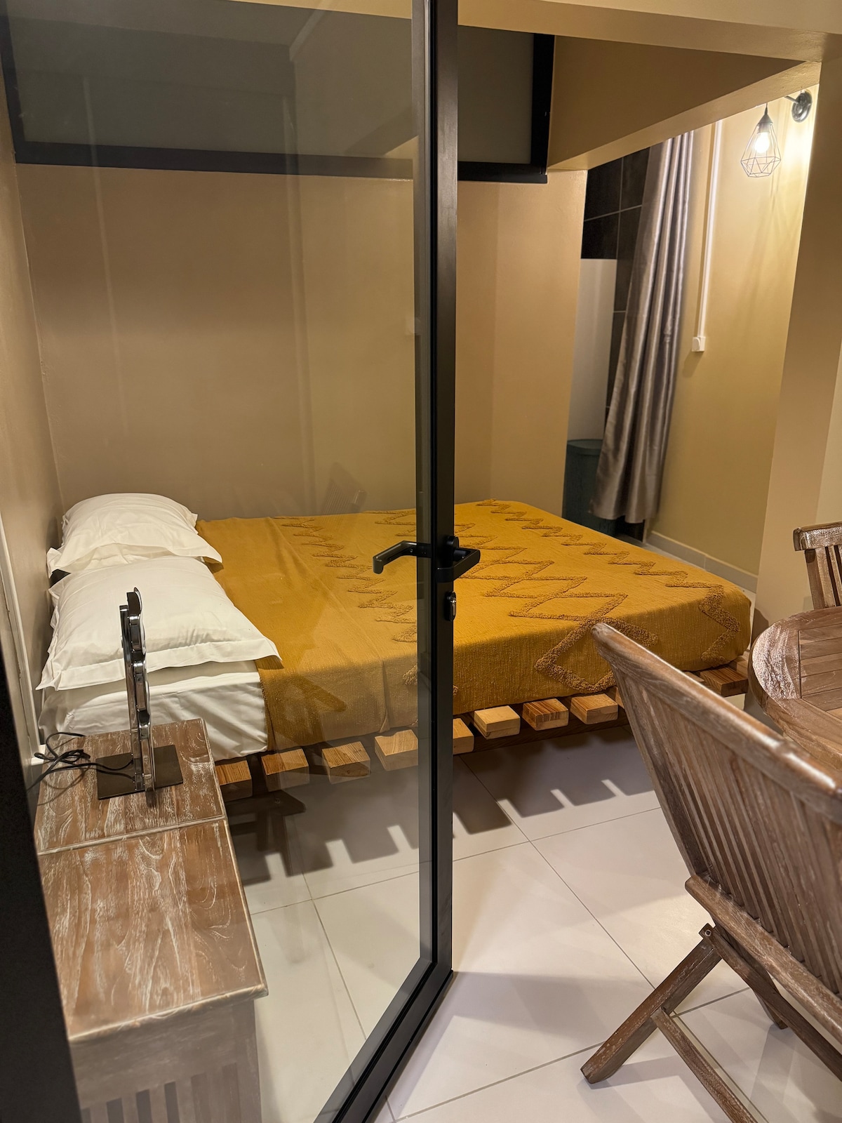 The image displays a cozy sleeping area separated by a glass partition. A spacious bed is dressed in a yellow blanket, complemented by crisp white pillows. A small wooden side table holds a TV, and a dining area is visible with wooden chairs in the foreground.