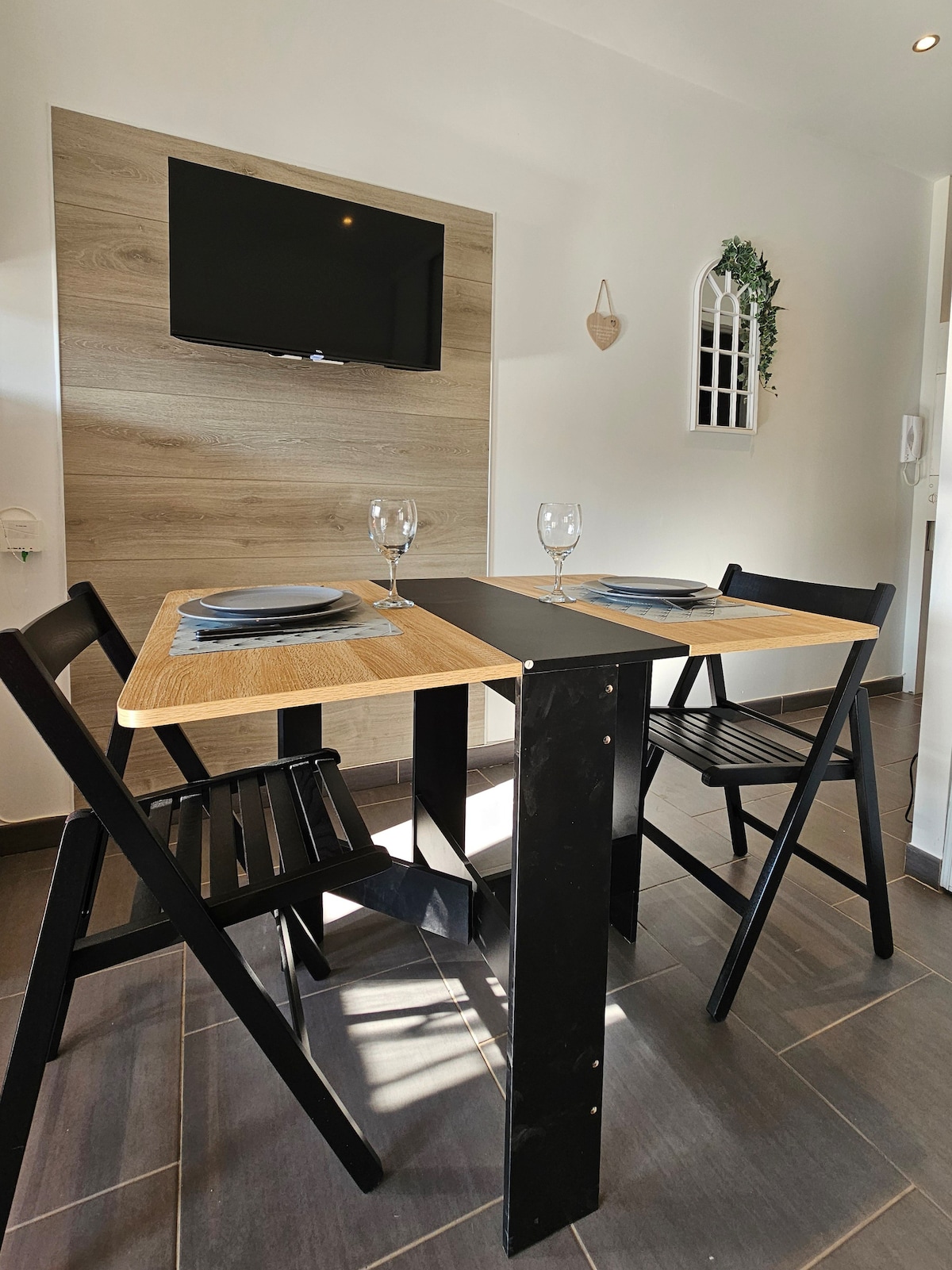 A dining area features a compact table with two chairs, suitable for enjoying meals. A flat-screen television is mounted on the wall, and a decorative arch window adds subtle charm to the space. Natural light enhances the warm tones of the surroundings.