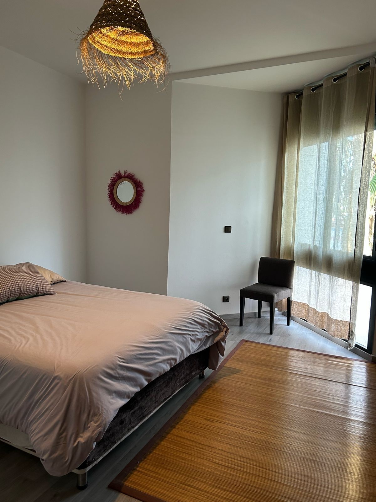 A well-lit bedroom features a spacious bed covered with a soft gray blanket, situated next to a large window draped with light, sheer curtains. A small round mirror is mounted on the wall, and a minimalistic chair is positioned in the corner.